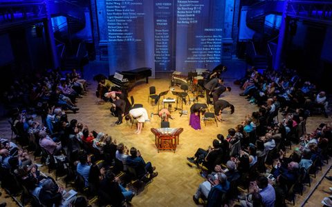Above the audience shot showing the Tangram collective bowing in the centre, surrounded by audience members on the outside. At the back, there are mesh screens with the performance credits projecting on them.