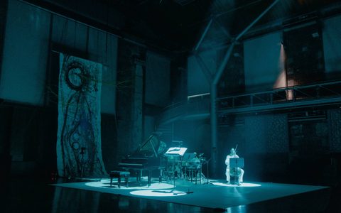 A zoomed out image of a cellist, in a spotlight. The stage has other instruments on it and in the background there is a large artwork with swirls and tree branches.