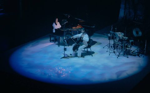 A blue circle spotlight shines upon a cellist wearing white and a pianist playing a grand piano. A drum kit is in the background.