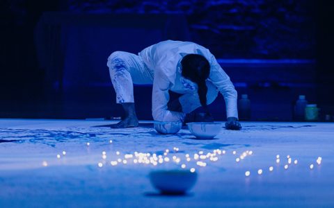 Echo Morgan, a performance artist, is crouched down on the floor with bowls of paint in front of her. Blue paint is scattered around and glowing balls of light are in the foreground.