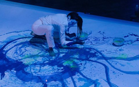 Echo Morgan, a performance artist, is crouched down on the floor with bowls of paint in front of her. Blue and green paint is scattered around a white background.