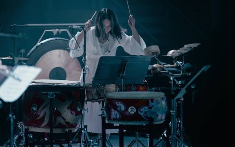 Beibei Wang is surrounded by percussion instruments, holding her sticks in the air.