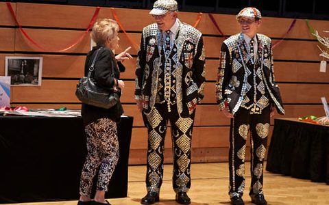 Two men standing wearing highly ornate and decorative suits with an elderly lady speaking to them on the left side