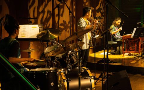 A person playing a drumkit is in the foreground with Daniel Shao playing the flute in the centre. Manta is in the background playing the yangqin.