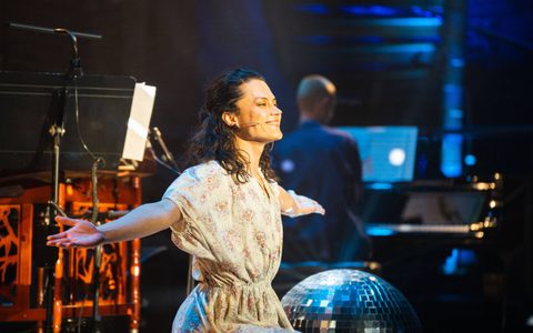 Manta wearing a floral dress smiling and holding their hands out wide. There is a glitter ball at the bottom of the image.