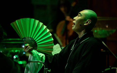 A bald Chinese man with a surprised expression, holding a fan, bathed in green light.