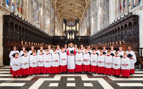 The members of The Choir of King's College, Cambridge, stand in the King's College Chapel, wearing white and red choir vestments. They stand in two symmetrical rows, with conductor Daniel Hyde standing in the center.