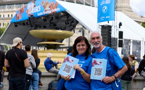 Two volunteers wearing blue t-shirts, smiling and holding plaquards with QR codes on them, stood in front of the BMW Classics stage.
