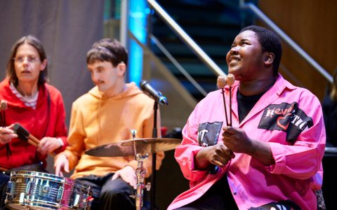 three people making music. One of them is smiling and holding mallets the other two are in front of a drum.