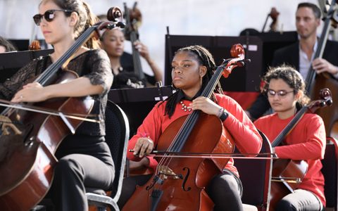 LSO Discovery participant wearing a red long sleeve shirt playing her cello performing with other LSO Discovery and LSO Musicians on the BMW Classics 2025 stage in Trafalgar Square, London