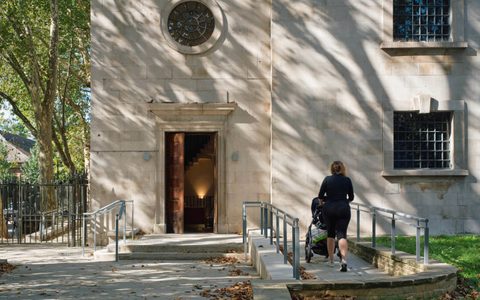 A person pushing a stroller towards the open door of a historic building on a sunny day, surrounded by trees and fallen leaves.