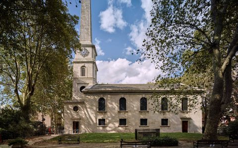 Exterior view of LSO St. Luke's in London, showing the church's classic architecture with a tall spire, surrounded by trees and benches under a clear sky.