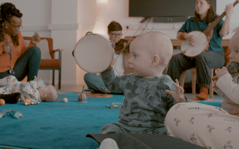 Two babies, one of them holding an instrument, parents and participants in the background.
