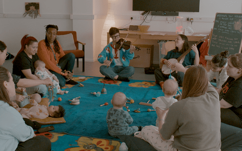 Parents with the babies sitting in a circle accompanied by a session leader and a musician, playing a violin.