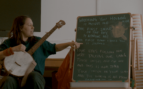 Instructor pointing on a board with the lyrics of a song written on it, while holding an instrument.